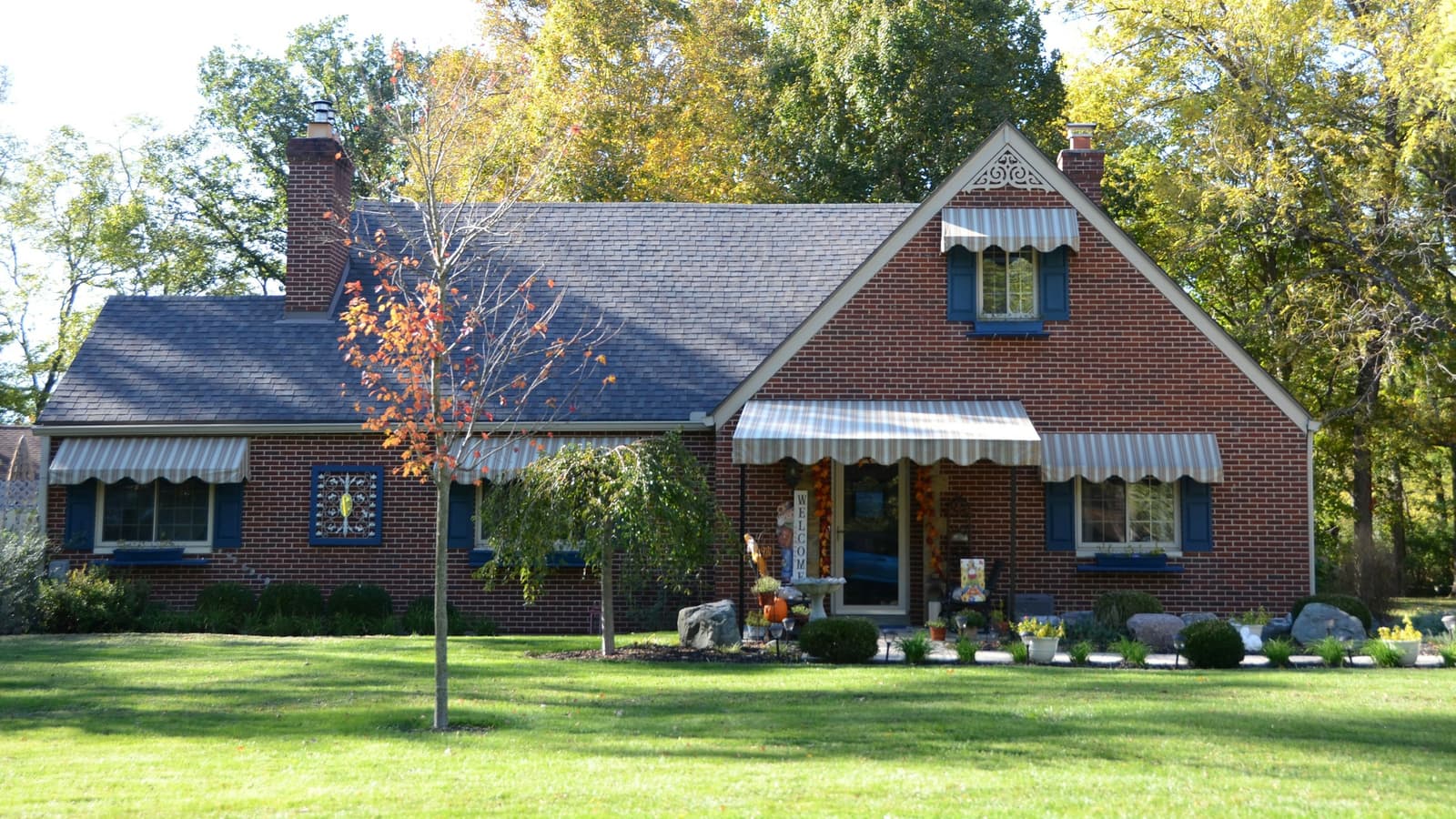 Brick cape-style house with striped awnings and a large manicured lawn in autumn